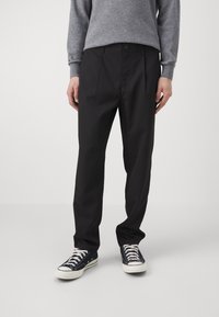 Osoba ubrana w czarne dopasowane spodnie, szary długi rękaw, oraz czarne niskie tenisówki z białymi podeszwami i sznurówkami, stojąca na białym tle.