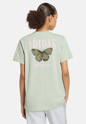 Kvinde iført en lysegrøn Adidas t-shirt med sommerfuglegrafik og teksten "sportswear" på ryggen, kombineret med lysegrå bukser.