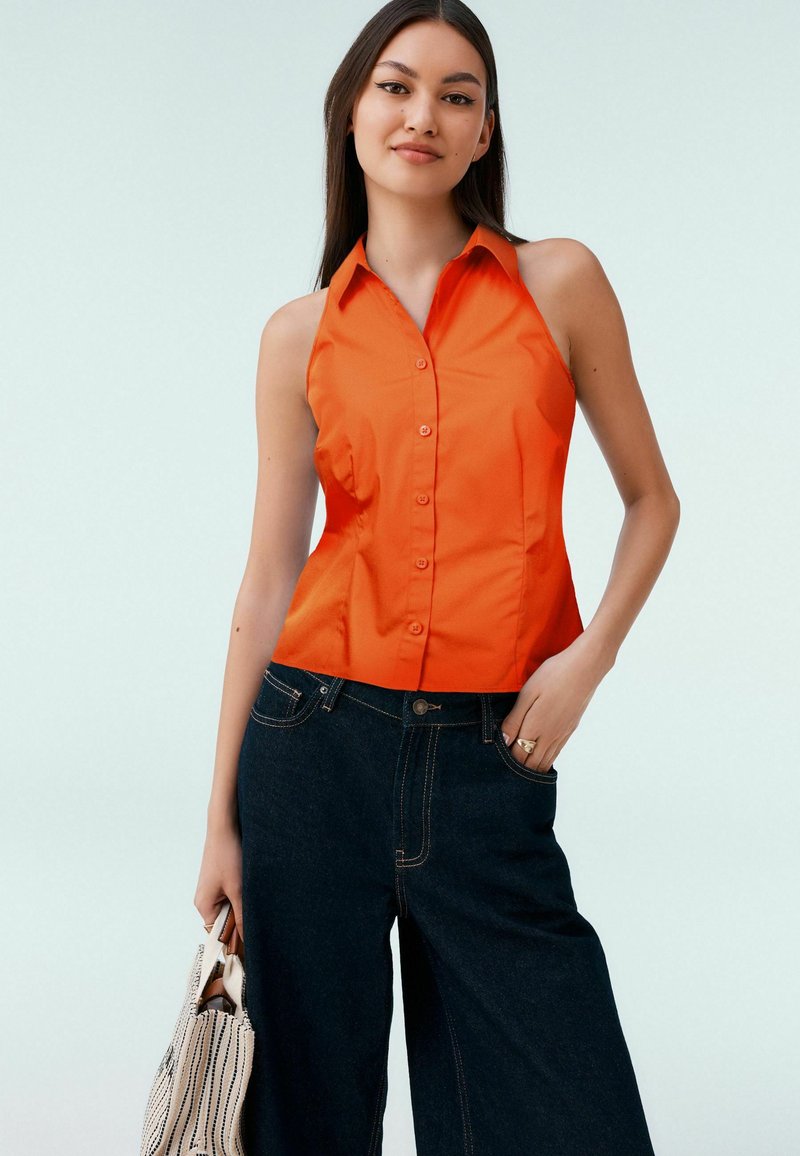 Haut sans manches orange à boutons avec col, design ajusté, texture lisse, associé à un jean en denim large de couleur sombre et un sac fourre-tout rayé.