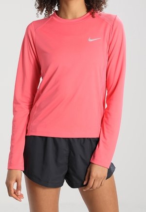 Vrouw draagt een koraalrode lange mouwen sporttop met een klein wit logo en zwarte hardloopshorts tegen een neutrale achtergrond.