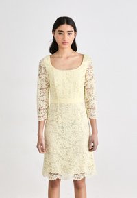 Robe en dentelle de couleur jaune clair avec un décollet�é carré, des manches en dentelle à trois quarts et un ourlet festonné ; texture de dentelle florale complexe sur toute la surface.