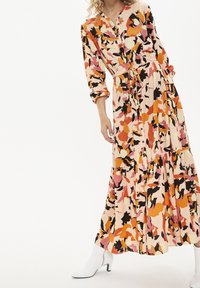 Bloemen-maxi-jurk in een perzikbasis met zwarte, oranje en roze patronen. Heeft een knoopsluiting aan de voorkant, lange mouwen en een geplooide rok.