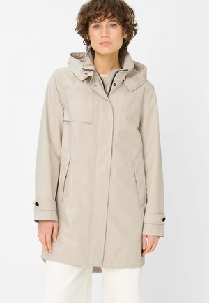Frau trägt eine beige Kapuzen-Regenjacke mit Druckknopfmanschetten, Reißverschluss und Druckknopfverschluss vorne, steht vor einem weißen Hintergrund.