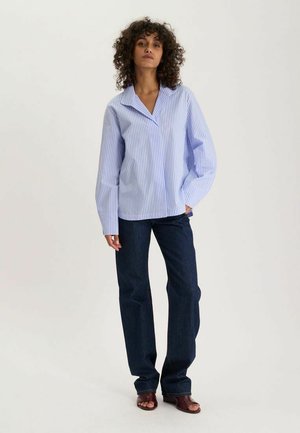 Femme aux cheveux bouclés portant une chemise à manches longues rayée bleu clair, un jean bleu foncé et des chaussures ouvertes marron, debout, une main dans la poche.