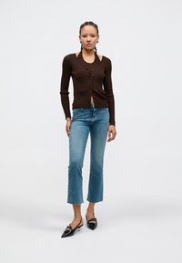 Καφέ ριμπ ζακέτα με V-λαιμόκοψη και κούμπωμα με κουμπιά, συνδυασμένη με ανοιχτό μπλε τζιν καμπάνα. Το μοντέλο φοράει μαύρα παπούτσια με μυτερή μύτη.