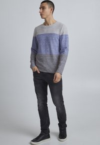 Svetra v sivi in modri barvi, blokirano po barvah, z okroglo izrezom, izdelan iz teksturirane pletene tkanine. V paru z črnimi prilegajočimi se jeans hlačami in črnimi supergami.