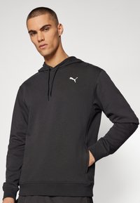 Ung mand med kort hår iført en sort Puma-hoodie med en frontlomme, stående foran en ensfarvet lys baggrund.