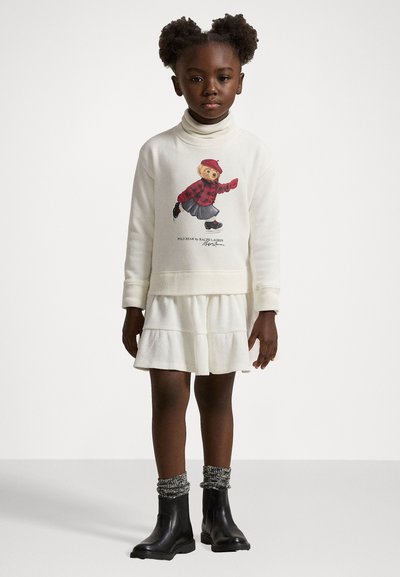 Baltā džemperkleita ar teddy lāča grafiku, garām piedurknēm un slāņotu svārku. Melni potīšu zābaki ar teksturētām zeķēm.