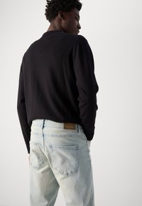 Black long-sleeve shirt paired with light blue jeans, featuring a distressed look and an INDICODE leather tag on the back waistband.