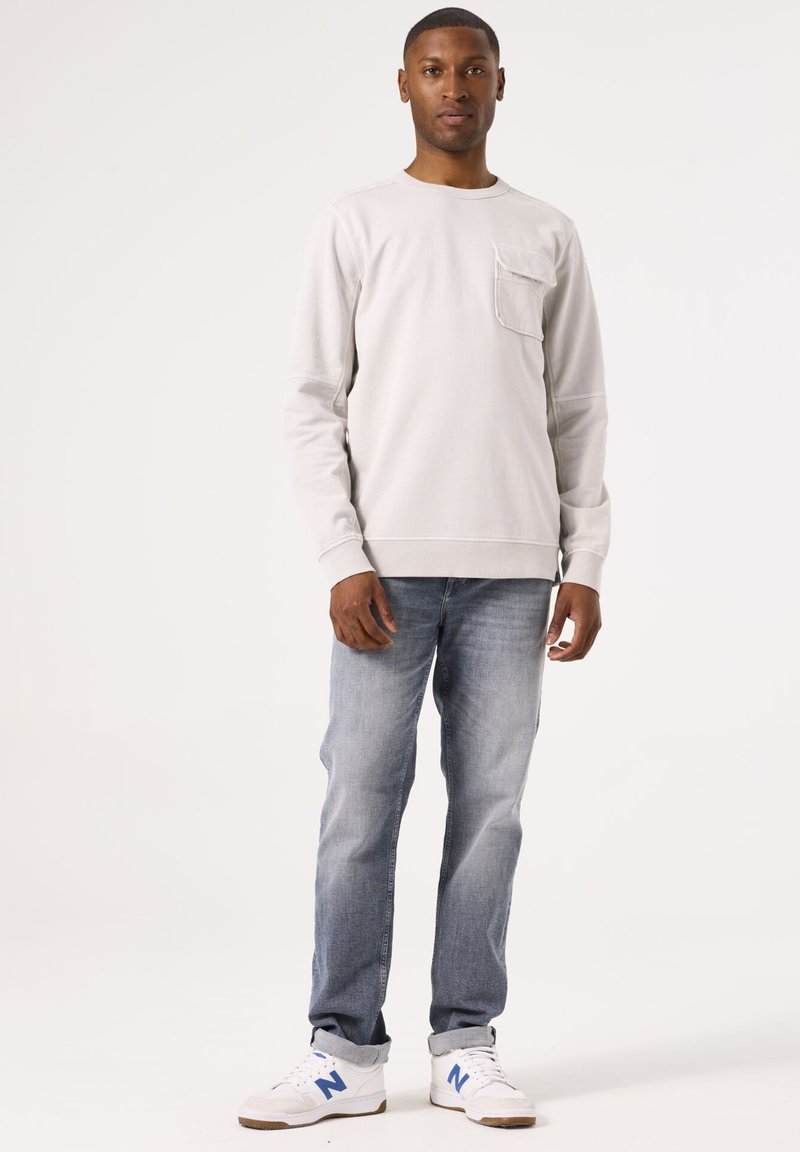 Sudadera de manga larga gris claro con un bolsillo en el pecho, combinada con jeans azules desgastados y zapatillas blancas con detalles en azul.