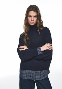 Granatowy sweter z golfem nałożony na niebieską, kraciastą koszulę, z luźnymi rękawami i widoczną fakturą, zestawiony z ciemnymi spodniami dżinsowymi.