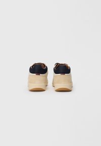 Tenisky s telovou semišovou štruktúrou a tmavomodrým koženým golierom. Majú podrážku z gumovej gumy a hnedé šnúrky na zadnej strane.