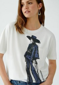 Weißes T-Shirt mit einer blauen und schwarzen grafischen Illustration einer Figur mit Hut und fransiger Kleidung. Kurze Ärmel und eine Taschenapplikation.