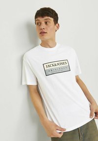 Balta kokvilnas T-krekls ar melnu un bežīgu taisnstūrveida logotipa druku un tekstu "JACK&JONES ORIGINALS", ar apaļu kaklu un īsām piedurknēm.