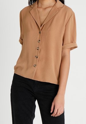 Vrouw die een lichtbruin overhemd met korte mouwen draagt, waarvan de mouwen zijn opgerold, en een zwarte broek, tegen een effen achtergrond.