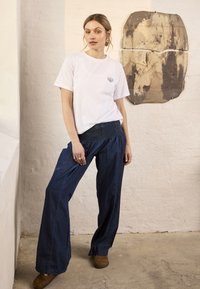 Femme portant une chemise blanche à manches courtes avec un petit emblème en coquillage et un pantalon large bleu marine, debout à l’intérieur contre un mur de couleur claire.