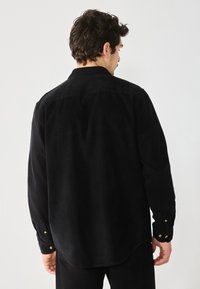 Black corduroy shirt with a back yoke, buttoned cuffs, and a slightly curved hem. Features a textured, soft fabric with visible wale pattern.