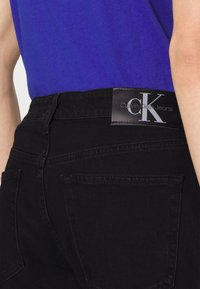 Osoba oblečená v černých džínách Calvin Klein s logem na pásku a s modrou košilí zastrčenou dovnitř.