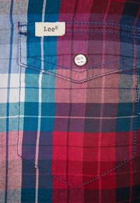 Red, blue, and white checkered fabric shirt featuring a pocket with a metal button, and a label reading "Lee®" stitched on the pocket.