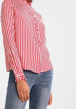 Rood-wit gestreept shirt met lange mouwen, verticale strepen, met ruches aan de knoopsluiting en manchetten, gedragen met een blauwe spijkerbroek door een staand persoon.