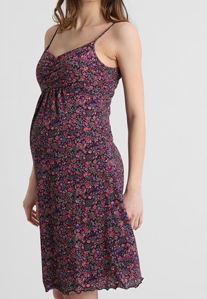 Zwangere vrouw in een mouwloze, knielange jurk met bloemenprint en dunne bandjes, staand tegen een effen lichtgrijze achtergrond.