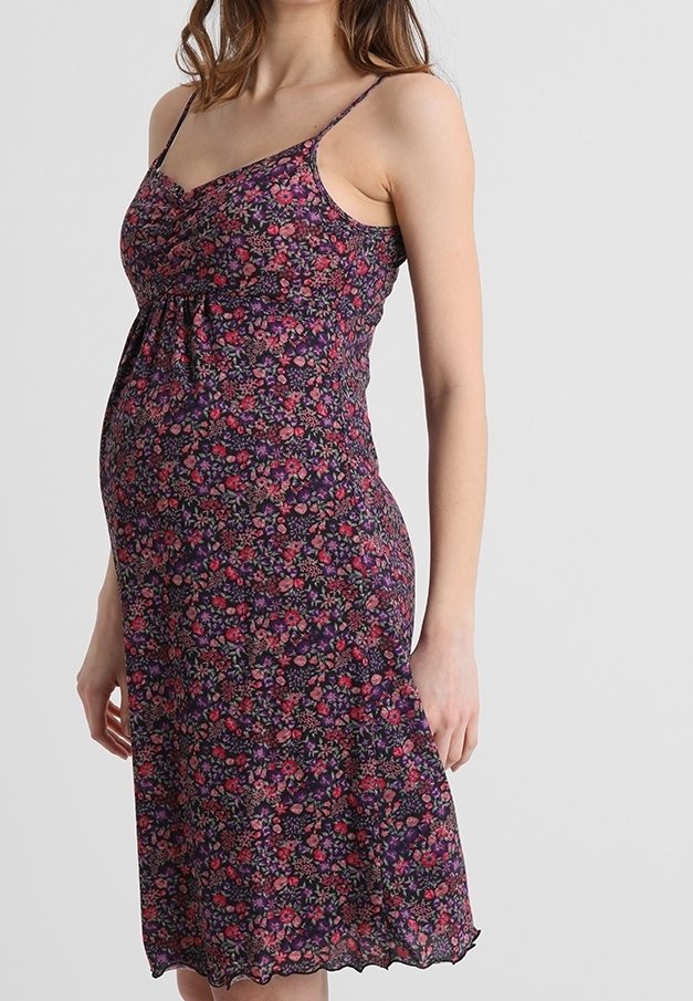 Femme enceinte portant une robe fleurie sans manches, arrivant au genou avec de fines bretelles, debout devant un mur gris clair uni.