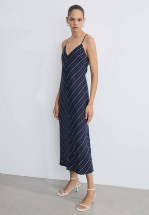 Donna in piedi in uno spazio minimalista indossa un abito midi senza maniche a V blu navy con sottili strisce diagonali bianche e sandali con tacco bianchi.