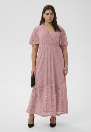 Nő, aki egy térdig érő, poros rózsaszín csipkeruhát visel fodros ujjakkal, kezében fekete borítéktáskával és fekete magassarkú cipőben áll, egyszínű háttér előtt.