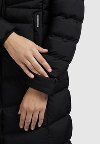 Mâini ajustând manșeta cu fermoar a unei jachete puffer negre cu eticheta "Khuloge Uninebrand" pe panoul pieptului.