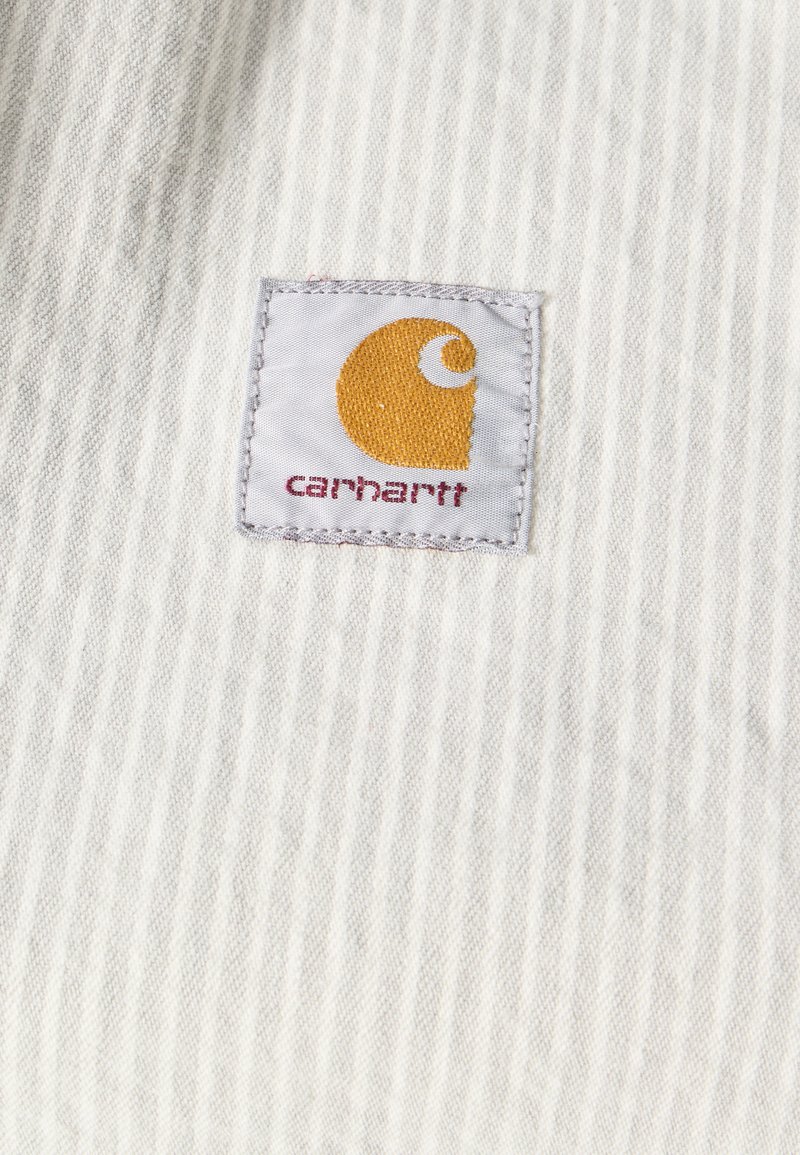 Witte, gestructureerde stof met een klein Carhartt-logoplaatje met een gele spiraal en rode tekst, gecentreerd op de stof.