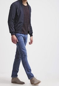Giacca bomber blu sopra un maglione marrone a trama, abbinata a jeans blu e stivaletti grigio chiaro con suola marrone.