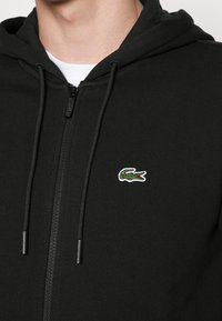 Zwarte zip-up hoodie van een katoenmix, met een groen-wit geborduurd krokodillenlogo op de borst en trekkoorden aan de capuchon.