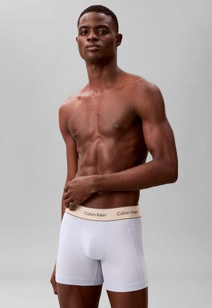 Muscular shirtless man wearing white Calvin Klein boxer briefs, standing against plain gray background with a neutral expression.