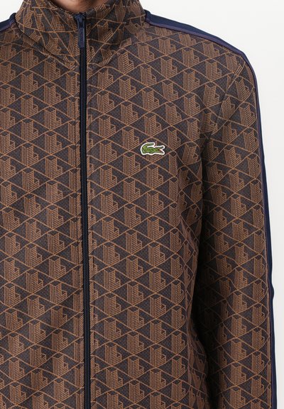 Veste zippée marron foncé avec des motifs géométriques orange. Dotée d'un col montant et d'un logo brodé en vert et blanc.