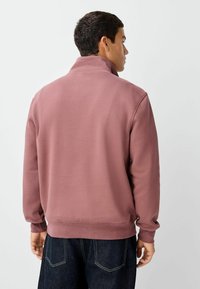 Sweter w kolorze mauve, wykonany z miękkiego materiału, z wysokim kołnierzem, długimi rękawami oraz prążkowanymi mankietami i dołem. Widoczne ciemnoniebieskie dżinsy poniżej.