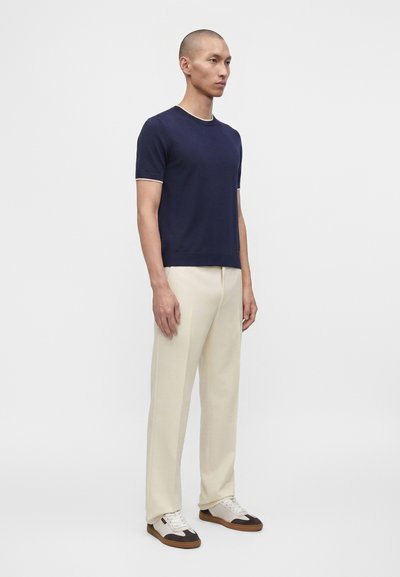 Homme debout portant une chemise bleu marine à manches courtes, un pantalon couleur crème et des baskets blanches avec des détails marron sur un fond clair uni.