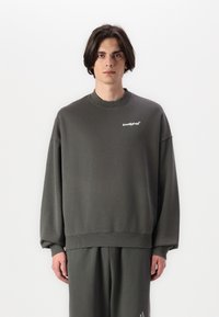 Per didelis tamsiai žalias džemperis iš minkštos medžiagos, turintis šonkaulinę apykaklę ir žemai nusileidusias rankoves, su baltu logotipu ant krūtinės.