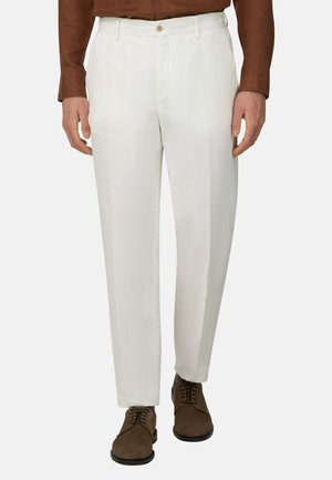 Homme portant un pantalon blanc sur mesure avec des plis à l'avant, une chemise marron à manches longues rentrée, et des chaussures marron foncé à lacets.