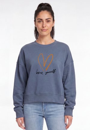 Femme aux cheveux foncés attachés portant un sweat bleu avec un cœur marron et le texte "love yourself", assorti à un jean bleu clair.
