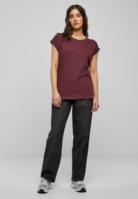 T-shirt bordeaux a maniche corte con vestibilità rilassata, abbinato a jeans dritti in denim scuro e scarpe da ginnastica grigie.