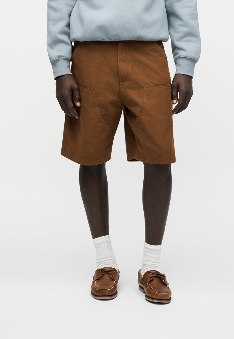 Uomo con pantaloncini marroni fino al ginocchio, calzini bianchi a metà polpaccio, scarpe da barca in pelle marrone e una felpa a manica lunga azzurra, in piedi.