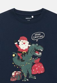 Marineblå t-skjorte med en grafikk av julenissen på en dinosaur, med fargerike julelys, en rød gavepose, og innpakkede gaver i bunnen.