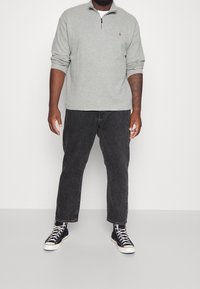 Man med grå kvartdraget sweatshirt, svarta urblekta jeans och svarta högsula kanvas sneakers som står mot en enkel vit bakgrund.