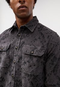 Chemise en velours côtelé gris avec un motif floral noir, dotée de deux poches poitrine et de fermetures à boutons, tissu texturé et col stylé.