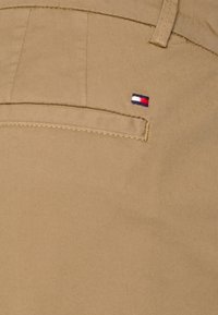 Pantaloni khaki con una tasca posteriore decorata da un piccolo logo ricamato in blu navy, rosso e bianco. Il tessuto ha una texture liscia e opaca.