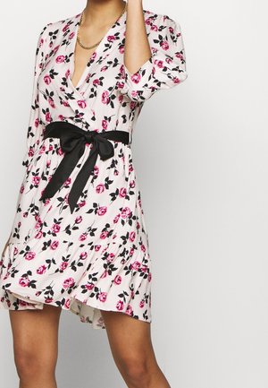 Vrouw met een witte bloemenjurk met roze rozen, zwarte bladeren, zwarte taille strik en gouden ketting, bij de schouders en halverwege de dijen bijgesneden.