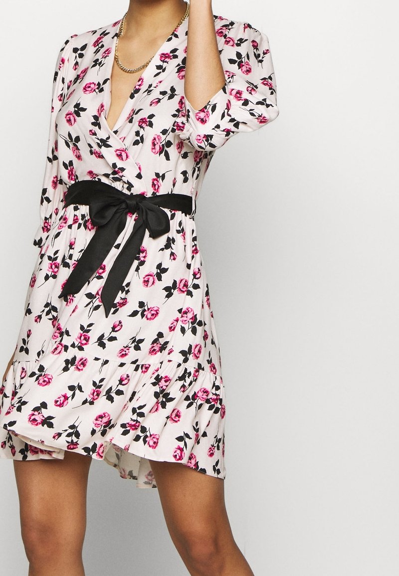 Femme portant une robe blanche à fleurs avec des roses roses, des feuilles noires, un nœud noir à la taille et un collier chaîne dorée, cadrée aux épaules et à mi-cuisse.