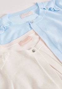 Két kardigán: az egyik világoskék, fodros díszítéssel és fényes gombbal, a másik krém színű, sima textúrával és hasonló gombtervezéssel.