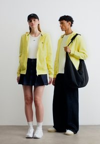 Deux personnes portant des sweats à capuche jaunes, des hauts blancs, des pantalons foncés et des chaussures blanches ; l'une d'elles tient un grand sac à bandoulière noir.