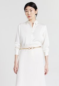 Maglietta bianca in satin con colletto, chiusura a bottoni e maniche lunghe, abbinata a una gonna coordinata e una cintura circolare dorata.